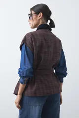 Chaleco de corte relajado con estampado a cuadros en tonos tierra, cuello camisero oscuro y cierre frontal con botones. Incluye un cinturón de la misma tela para ajustar a la cintura y bolsillos frontales.