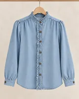 Camisa de denim de algodón celeste, con cuello alto con volados, botonadura frontal oculta con detalles de volados, mangas largas abullonadas con puños ajustados con botones y frunces en los hombros.