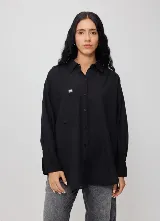 Camisa negra de manga larga con cierre frontal de botones, cuello camisero y bolsillo en el pecho con pequeño parche bordado de la marca. Corte holgado y relajado.