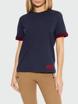 Remera Moncler de algodón azul marino con cuello redondo y manga corta. Presenta puños tejidos con patrón en zigzag rojo y azul marino, y un parche rectangular rojo con el logo de la marca en el borde inferior.