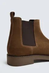 Bota estilo Chelsea de hombre, color marrón, fabricada en piel con acabado serraje (gamuza). Presenta elásticos laterales oscuros y suela de goma a tono con tacón bajo.