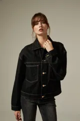 Campera de jean color negro con costuras a contratono, diseño de corte holgado, cuello camisero, cierre frontal con botones metálicos y dos bolsillos frontales con solapa.