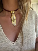 Gargantilla con cinta de terciopelo color borgoña y dije colgante de metal dorado con forma de hoja perforada y detalles plateados.