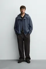 Parka de tejido técnico con acabado engomado, cuello subido y capucha ajustable. Presenta cierre frontal con cremallera oculta bajo solapa con botones a presión, bolsillos laterales de vivo y mangas largas con puños ajustables.