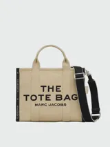 Bolso tipo tote bag de Marc Jacobs en color beige, confeccionado en lona resistente. Presenta el texto "THE TOTE BAG" y "MARC JACOBS" impreso en la parte frontal en color negro. Cuenta con asas superiores y una correa de hombro ajustable y extraíble, además de ribetes laterales negros con inscripciones.