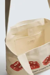 Bolso tipo shopper de tela color beige, con asas cortas y asa de hombro. Estampado frontal con cuatro dados rojos con corazones y texto que dice "SOMETIMES SÍ, SOMETIMES BYE".