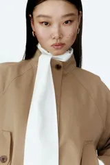 Campera oversized de sarga de algodón color beige, con cuello elevado, cierre frontal oculto y solapa rompevientos. Cuenta con mangas raglán, bolsillos delanteros con solapa y botón, y cordón ajustable en el bajo que frunce la cintura.