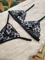 Conjunto de lencería compuesto por un bralette triangular y una bombacha tipo tanga, confeccionados en encaje negro con detalles florales en hilo plateado.