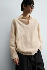 Camisa de manga larga color beige claro con textura jacquard arrugada, cuello solapa con abertura en V y bolsillo canguro frontal. Posee cordones ajustables en el bajo lateral.