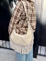 Cartera tipo hobo de cuero color arena, con diseño de media luna y flecos largos en la parte inferior. Cuenta con correa ajustable para llevar al hombro y cierre superior.