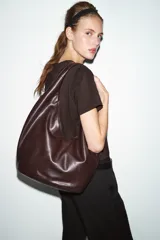 Bolso de hombro tipo saca en formato maxi, confeccionado en material sintético negro. Presenta un diseño holgado con asa integrada, cierre imantado en el compartimento principal, bolsillo lateral con cremallera y detalle de tirador.