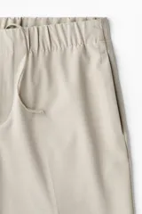 Pantalón de corte relajado color beige claro, con cintura elástica ajustable con cordón oculto y pliegues frontales. Presenta bolsillos laterales y bolsillos traseros ribeteados.
