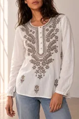 Blusa blanca de manga larga con detalles bordados en tono marrón en el pecho y las mangas, cuello redondo y cierre frontal con pequeños botones.