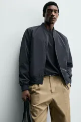 Cazadora bomber ligera de tejido técnico color verde oliva, con cuello, puños y bajo en rib elástico. Presenta cierre frontal de cremallera y bolsillos de vivo en la cadera.