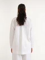 Camisa blanca de manga larga con cuello clásico y cierre frontal de botones, de corte recto y largo por debajo de la cadera.