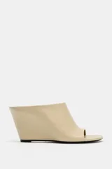 Sandalia tipo mule con tacón cuña, confeccionada en piel color beige. Presenta diseño destalonado, apertura frontal y punta cuadrada asimétrica.
