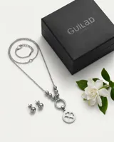 Set de joyería en acero compuesto por un collar con dije circular de trébol de cuatro hojas y un par de aros tipo bolita. El collar presenta una cadena con esferas decorativas en la parte central.