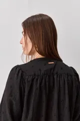Camisa negra de textura liviana con detalle de lunares en relieve, cuello mao y cierre frontal con botones. Presenta mangas 3/4 con puños elastizados y frunces en los hombros.