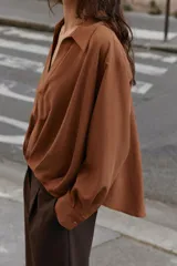Camisa de lino color beige, con cuello solapa, escote en V y mangas largas. Presenta un diseño de bajo asimétrico con pliegues delanteros que generan un efecto abullonado.