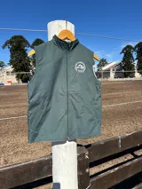 Chaleco verde oscuro de microfibra con cuello alto, cierre frontal y bolsillos laterales. Presenta un bordado circular con la silueta de un caballo en el pecho.