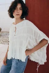 Top blanco de cuello redondo y mangas tipo capa, con textura fruncida, bajo asimétrico, lazos decorativos en el escote y en los laterales, y cierre posterior con botón.