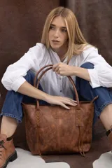 Cartera tipo tote bag de cuero vacuno marrón, con detalles de trenzado artesanal en los laterales y flecos colgantes decorativos. Cuenta con doble asa de hombro y cierre superior.