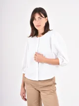 Camisa blanca de corte clásico con cuello redondo y cierre frontal oculto. Presenta mangas tres cuartos abullonadas con puños elásticos, brindando un toque femenino y moderno.