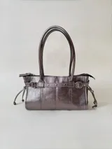 Cartera de cuero de tamaño medio con estructura rígida. Presenta una tira decorativa con tachas y hebillas metálicas en los extremos, además de lazos laterales. Cuenta con doble asa para llevar al hombro y cierre superior.