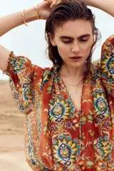 Vestido túnica corto y oversized de tejido plano con estampado tipo paisley y floral en tonos naranjas y azules. Presenta cuello redondo fruncido con abertura en V y lazos finos al frente, mangas cortas abullonadas con puños elásticos, cintura fruncida y falda con vuelo.