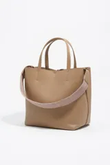 Bolso de hombro tipo tote bag de piel vacuna color beige, con doble asa de mano y correa ajustable de tejido. Presenta un cierre de cordón en la parte superior y un logo metálico en relieve en el frente. Incluye bolsillo interior con cierre de cremallera.