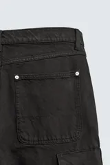 Pantalón cargo de corte relajado en color negro con efecto lavado. Presenta bolsillos laterales tipo cargo con solapas y cintas ajustables, bolsillos frontales y traseros, y detalles de argollas metálicas. Cierre frontal con cremallera y botón.