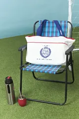 Bolso tipo shopper color blanco con detalles en rojo, confeccionado en algodón. Presenta un estampado central en azul con el escudo nacional y la frase 'El País de los mates y las siestas'. Cuenta con correas de hombro en color azul.