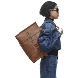 Bolso tipo tote de gran tamaño, confeccionado en cuero marrón con textura granulada. Presenta el logo de la marca estampado en el frente con la inscripción The Tote Bag Marc Jacobs.