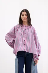 Camisa de corte holgado con diseño de rayas verticales en tonos fucsia, blanco y azul. Presenta cuello redondo con volado, cierre frontal con botones azules y mangas largas abullonadas con hombros caídos.