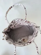 Cartera de cuero de tamaño medio con estructura rígida. Presenta una tira decorativa con tachas y hebillas metálicas en los extremos, además de lazos laterales. Cuenta con doble asa para llevar al hombro y cierre superior.