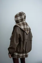 Chaqueta de cuero sintético color marrón con capucha y solapas amplias en tela escocesa a cuadros. Posee cierre frontal, puños elásticos y diseño de corte holgado.