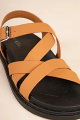 Sandalia baja de tiras cruzadas color camel, con hebilla metálica ajustable en el tobillo y suela negra gruesa.
