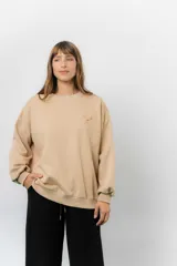 Buzo oversize de algodón frizado color beige, con cuello redondo, puños y cintura tejidos. Presenta un bordado pequeño de Snoopy en el frente.