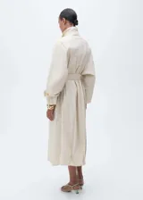 Abrigo largo de diseño oversize en color beige, confeccionado en tejido de algodón con elastano. Presenta cuello alto abotonado, cierre frontal con botones, mangas largas con trabillas en los puños y cinturón tipo batín para ajustar a la cintura. Incluye dos bolsillos laterales.