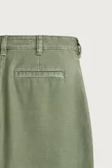 Pantalón chino baggy fit color verde militar, confeccionado en denim de algodón rígido, con corte holgado, tiro medio y pliegues frontales. Presenta bolsillos laterales y cierre frontal con cremallera y botón, con efecto lavado.