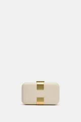 Bolso tipo clutch rígido de mano, color beige, con detalle de apliques metálicos dorados en el frente y cierre mediante clip. Incluye cadena extraíble para usar como bandolera.