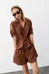 Chaqueta estilo safari de color marrón, con escote en pico, manga corta con cordones ajustables con abalorios y bolsillos delanteros de plastrón. Cuenta con cordón ajustable en la cintura y cierre frontal con botones.