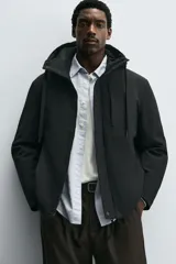 Cazadora técnica regular fit de color negro, con interior ligeramente acolchado. Presenta cuello subido con capucha ajustable, cierre frontal de cremallera oculta por solapa con botones a presión, bolsillos laterales y puños con interior de rib.