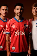 Camiseta de fútbol Umbro del Club Nacional de Football, modelo Away 2025 de color rojo con detalles en azul en el cuello en V y en las mangas. Cuenta con patrocinadores impresos en blanco, el logo de la marca y el escudo del club.