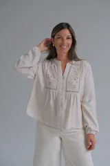 Blusa de color beige con cuello mao y cierre frontal de botones. Presenta detalles de bordado floral y calados en el frente, con mangas largas de corte holgado.
