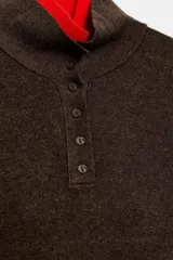 Jersey de punto con efecto doble, cuello subido y manga larga. Presenta un diseño en color marrón con detalles en rojo vibrante en el cuello, puños y ruedo. Cuenta con cierre frontal parcial mediante botones.
