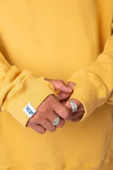 Buzo canguro amarillo ocre oversize de algodón rústico con capucha, cordones negros con letras blancas, bolsillo frontal y puños y cintura acanalados. Presenta estampados en el frente y las mangas.