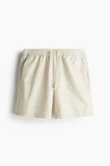 Short de lona de algodón color beige claro, con cintura elástica ajustable mediante cordón, bolsillos laterales al bies y un bolsillo insertado trasero. Presenta un cierre frontal decorativo.