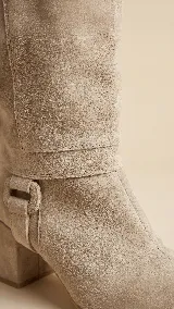 Botas de caña alta desmontables de gamuza color beige, con taco cuadrado de 6 cm y punta semi cuadrada. Presentan un diseño con costura central y detalle de estribo con hebilla forrada en el tobillo.