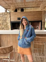 Campera de jean azul con capucha y detalles de bordado artesanal multicolor en la capucha, el pecho, los bolsillos y las mangas. Cuenta con cierre frontal de botones, bolsillos delanteros y diseño cómodo.
